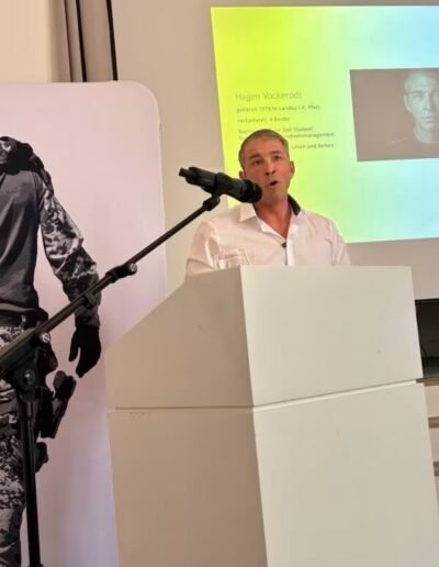 Veteranenwochenende 2024 mit dem 8.Tag der EinsatzVeteranen 6 Mann am Rednerpult beim Bund Deutscher Veteranen. Präsentation mit Porträt von Hagen Vockeroidt, Figur in Tarnkleidung links.