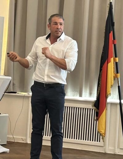 Veteranenwochenende 2024 mit dem 8.Tag der EinsatzVeteranen 7 Ein Mann hält beim Bund Deutscher Veteranen eine Präsentation, steht neben deutscher Flagge vor beigen Vorhängen.