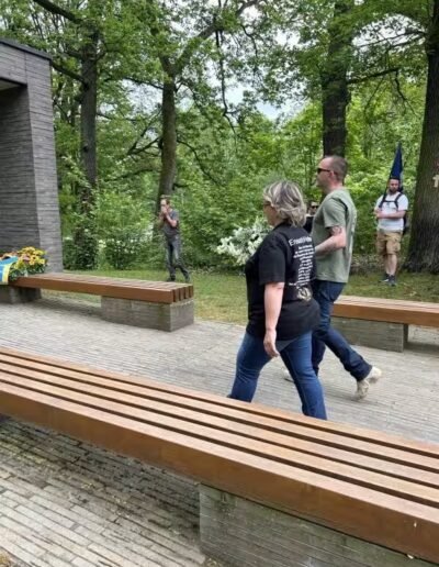 Veteranenwochenende 2024 mit dem 8.Tag der EinsatzVeteranen 36 Zwei Menschen mit weißen Blumen gehen auf einem Weg zum Veteranendenkmal, umgeben von Bäumen, Fahne und Fotograf im Hintergrund.