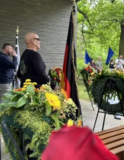Veteranenwochenende 2024 mit dem 8.Tag der EinsatzVeteranen 49 Mann in Schwarz steht bei Blumenarrangement am Gedenkort, mit Kränzen und Flaggen vom Bund Deutscher Veteranen im Hintergrund.