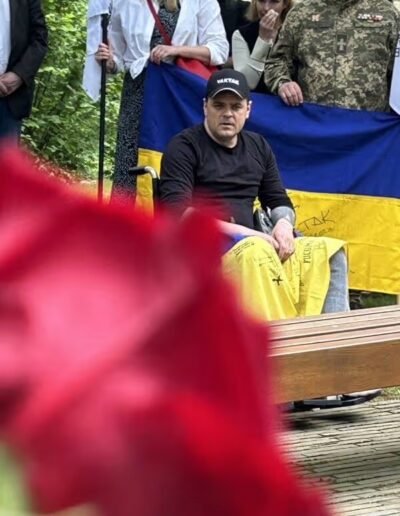 Veteranenwochenende 2024 mit dem 8.Tag der EinsatzVeteranen 64 Rollstuhlfahrer mit schwarzer Kleidung vor ukrainischer Flagge, mehrere Personen dahinter, rote Blume im Vordergrund unscharf.
