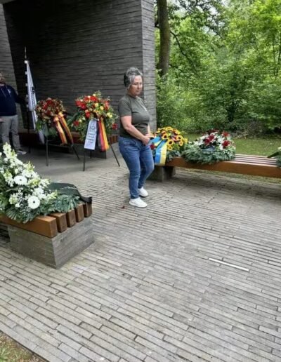 Veteranenwochenende 2024 mit dem 8.Tag der EinsatzVeteranen 63 Frau am Mahnmal des Bund Deutscher Veteranen, bunte Kränze und Blumen auf Bänken, umgeben von viel Grün.
