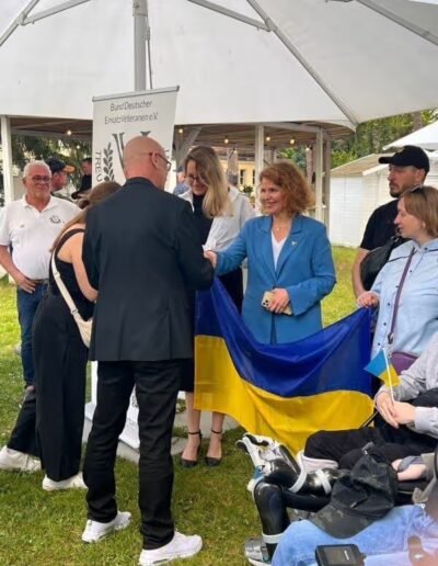 Veteranenwochenende 2024 mit dem 8.Tag der EinsatzVeteranen 15 Menschen versammeln sich mit ukrainischer Flagge, Frau im blauen Blazer begrüßt Mann. Auch Rollstuhlfahrer dabei.