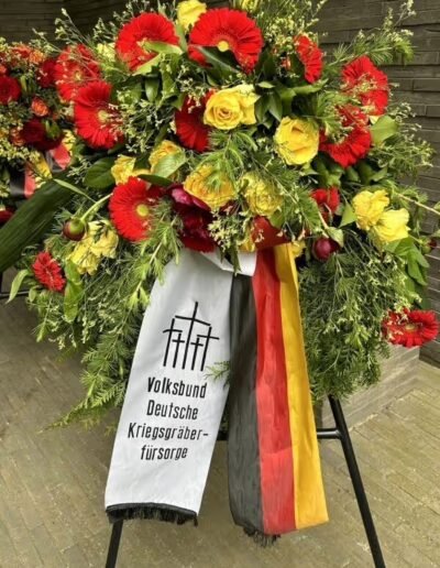 Veteranenwochenende 2024 mit dem 8.Tag der EinsatzVeteranen 76 Blumenkranz in Rot-Gelb mit Deutschlandband und Volksbund-Schleife vor Backsteinwand, Thema Bund Deutscher Veteranen.