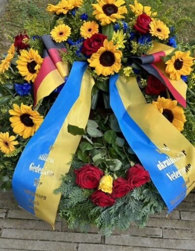 Veteranenwochenende 2024 mit dem 8.Tag der EinsatzVeteranen 75 Kranz mit Sonnenblumen, roten Rosen und blauem Blumenschmuck, Schleifen in Deutschlandfarben, draußen auf Stein abgelegt.