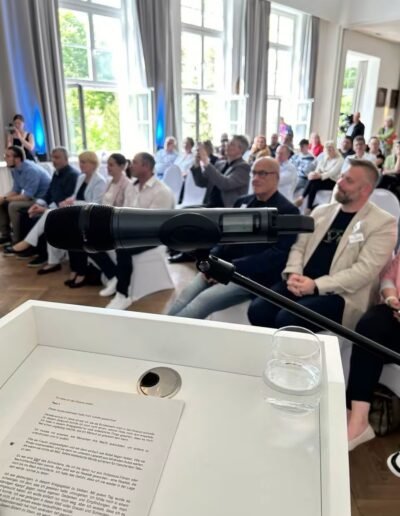 Veteranenwochenende 2024 mit dem 8.Tag der EinsatzVeteranen 2 Podium im hellen Saal mit Mikrofon und Wasserglas, Publikum verfolgt aufmerksam eine Veranstaltung des Bund Deutscher Veteranen.