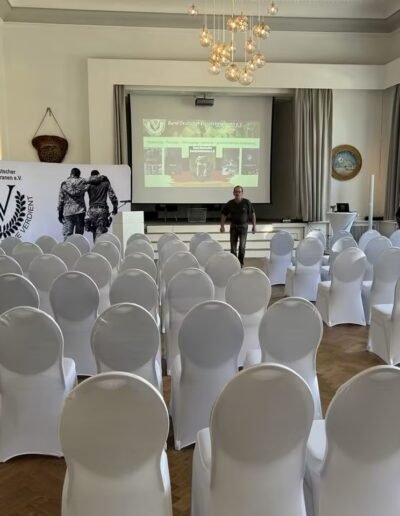 Veteranenwochenende 2024 mit dem 8.Tag der EinsatzVeteranen 1 Konferenzraum des Bund Deutscher Veteranen mit Stuhlreihen, Bühne, Beamer und zwei Personen, Sonnenlicht durch Fenster.
