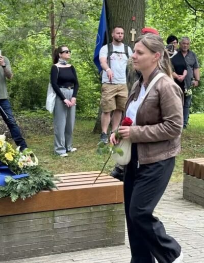 Veteranenwochenende 2024 mit dem 8.Tag der EinsatzVeteranen 54 Frau mit Rose und Kerze geht am Mahnmal mit Blumen; Menschen stehen im Freien bei Bäumen. Thema: Bund Deutscher Veteranen.