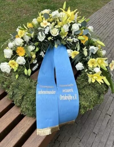 Veteranenwochenende 2024 mit dem 8.Tag der EinsatzVeteranen 71 Blumengesteck in Weiß und Gelb mit blauer Schleife "Bund Deutscher Veteranen" auf Holzbank neben Weg im Freien.