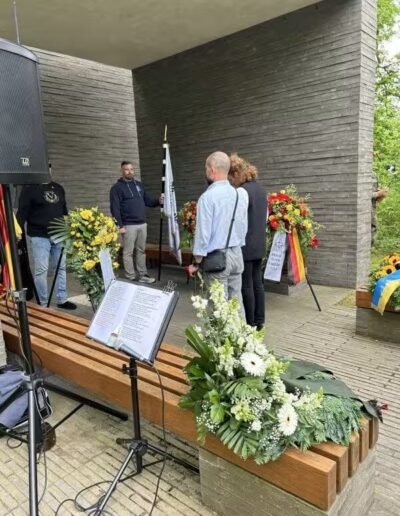 Veteranenwochenende 2024 mit dem 8.Tag der EinsatzVeteranen 53 Gedenkfeier des Bund Deutscher Veteranen: Menschen stehen vor Blumenkränzen an einem Memorial mit Musikständern und Lautsprecher.