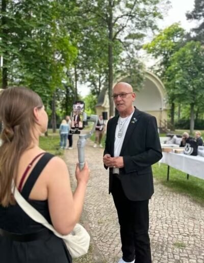 Veteranenwochenende 2024 mit dem 8.Tag der EinsatzVeteranen 18 Eine Frau filmt einen Mann im schwarzen Anzug im Park für den Bund Deutscher Veteranen, mit Bäumen und Tischen im Hintergrund.