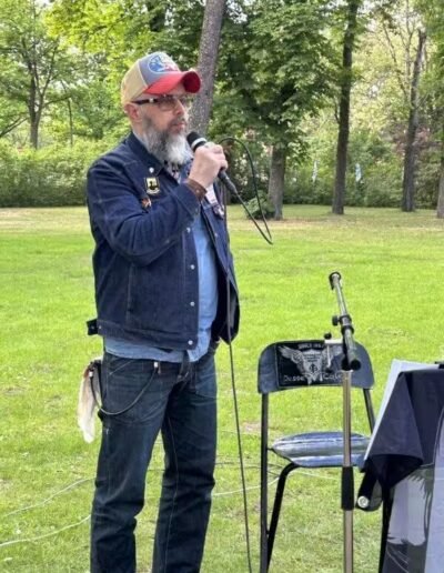 Veteranenwochenende 2024 mit dem 8.Tag der EinsatzVeteranen 21 Ein bärtiger Mann spricht beim Bund Deutscher Veteranen draußen ins Mikrofon, umgeben von Stuhl, Notenständer und Bäumen.