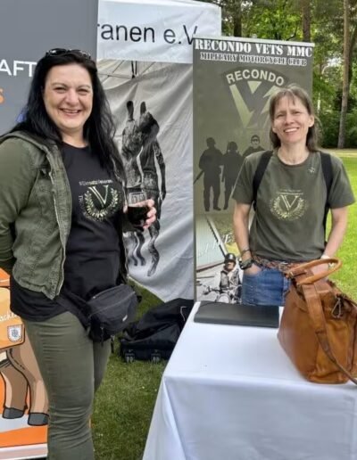 Veteranenwochenende 2024 mit dem 8.Tag der EinsatzVeteranen 20 Zwei Frauen am Infostand des Bund Deutscher Veteranen im Freien, tragen Recondo Vets Shirts, mit Bannern und Natur im Hintergrund.