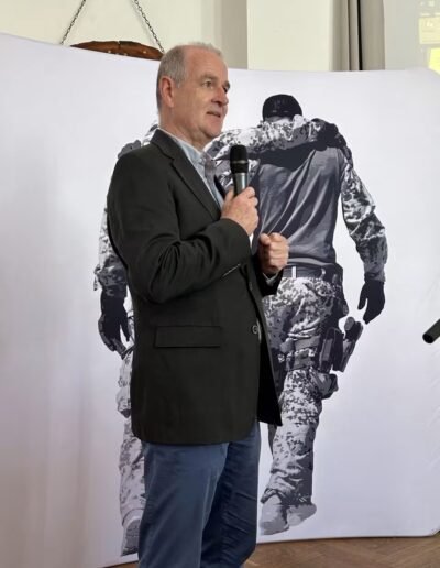Veteranenwochenende 2024 mit dem 8.Tag der EinsatzVeteranen 3 Mann im Blazer spricht mit Mikrofon vor großem Hintergrundbild von zwei Bundeswehrsoldaten, einer stützt den anderen.