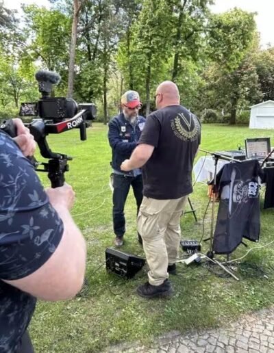 Veteranenwochenende 2024 mit dem 8.Tag der EinsatzVeteranen 22 Im Park sprechen zwei Männer neben Technik und Fahnen, ein Kameramann filmt. Thema: Bund Deutscher Veteranen, grüne Kulisse.