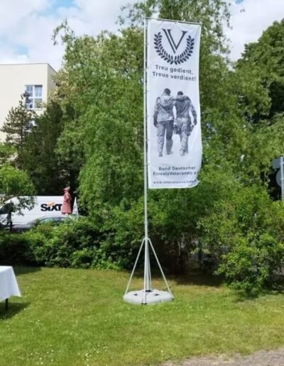 Veteranenwochenende 2024 mit dem 8.Tag der EinsatzVeteranen 11 Zwei Veteranen in Uniform gehen auf Banner Arm in Arm, großes V mit Lorbeerkranz, Schriftzug: Treu gedient, Treue verdient!.