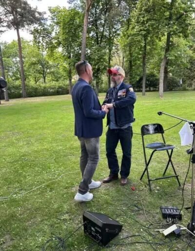 Veteranenwochenende 2024 mit dem 8.Tag der EinsatzVeteranen 23 Zwei Männer unterhalten sich im Park, umgeben von Bäumen. Einer hält eine Gitarre, Musik-Equipment und Stuhl stehen daneben.